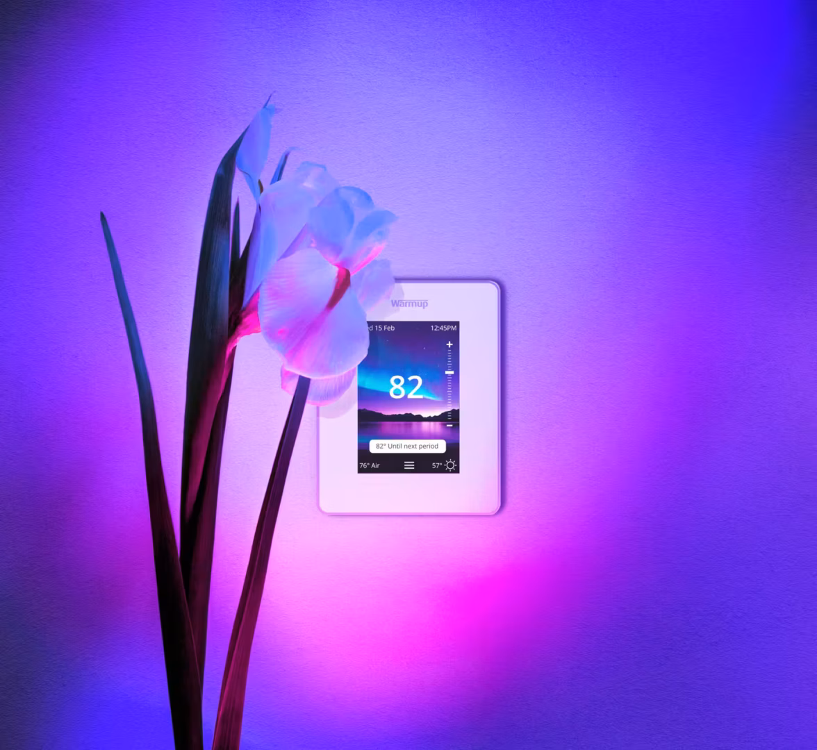 Warmup's 7iE Smart Matter Thermostat mounted on the wall next to an orchid plant.